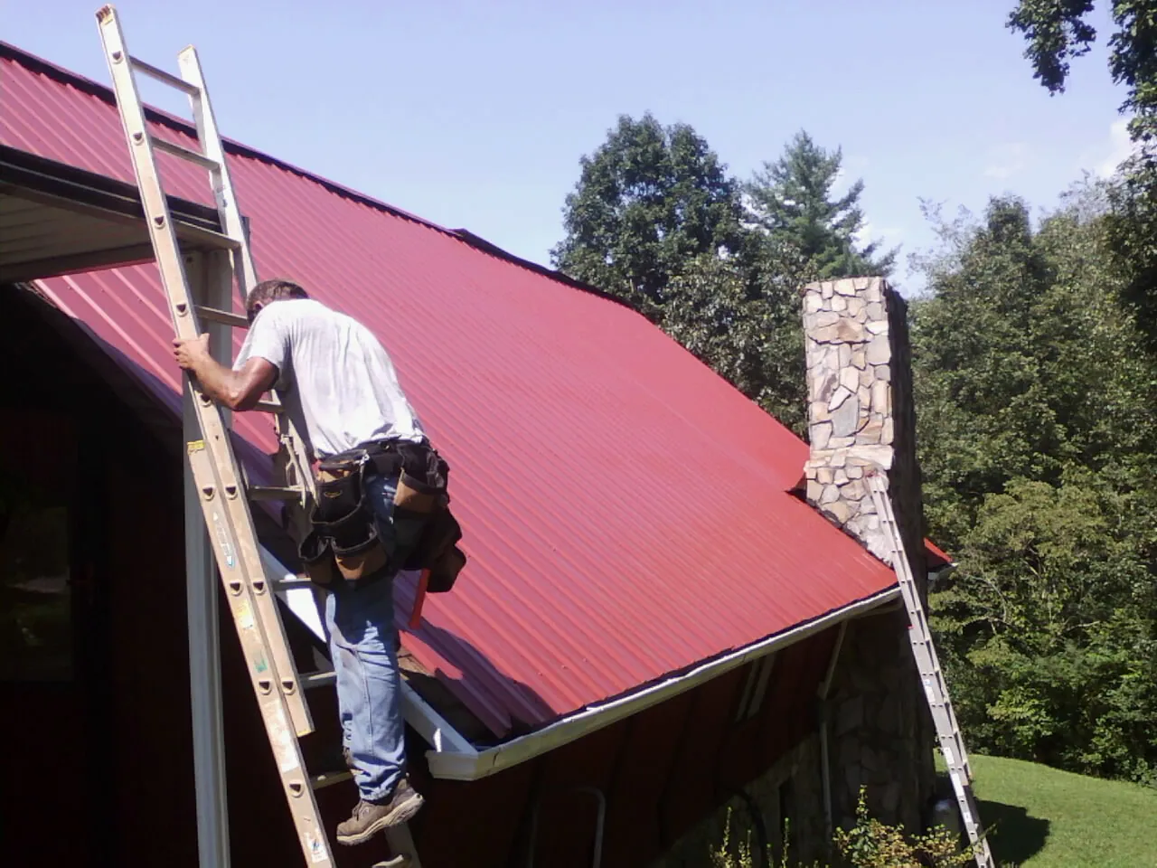Roofer on a ladder beside a red metal roof and stone chimney — Soffit & Fascia Repair by Roofing Brothers in Augusta